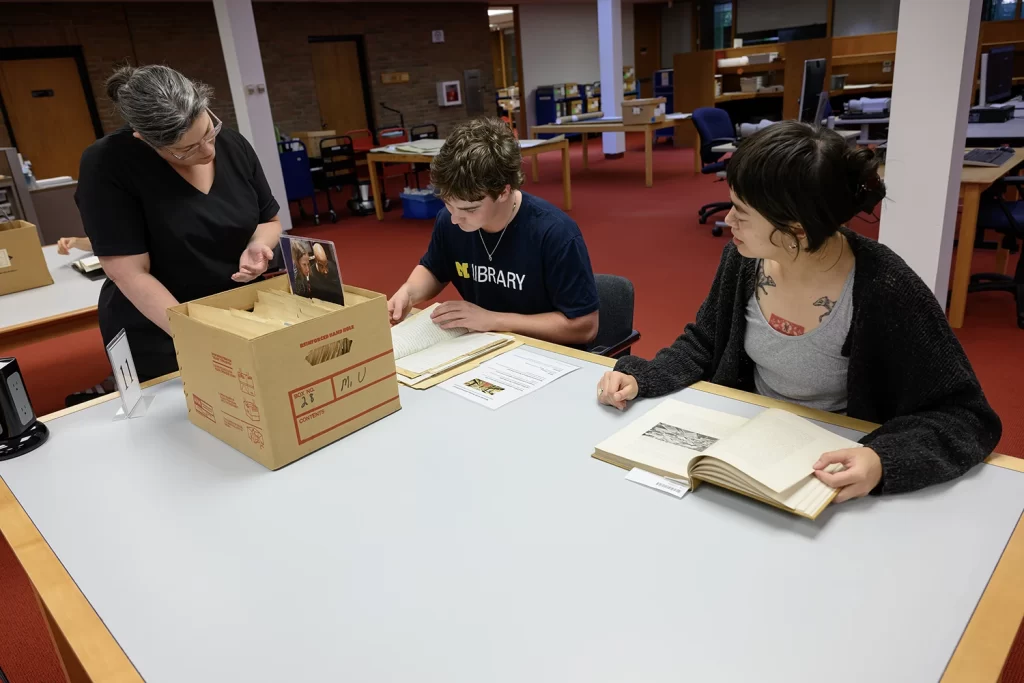 Two researchers, a man and a woman, sit at a table in the Bentley Historical Library's reading room. To the left in the photo is an archivist who is wearing a black dress and leaned over slightly to help them look at historical materials.