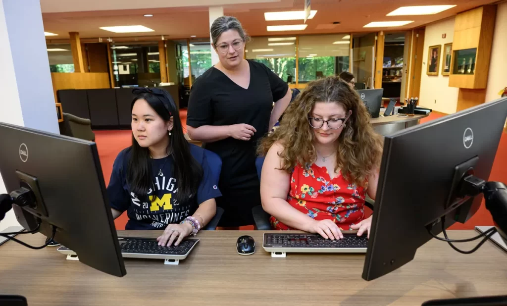 Two women sit at computer monitors with keyboards while a third woman stands behind them to help.