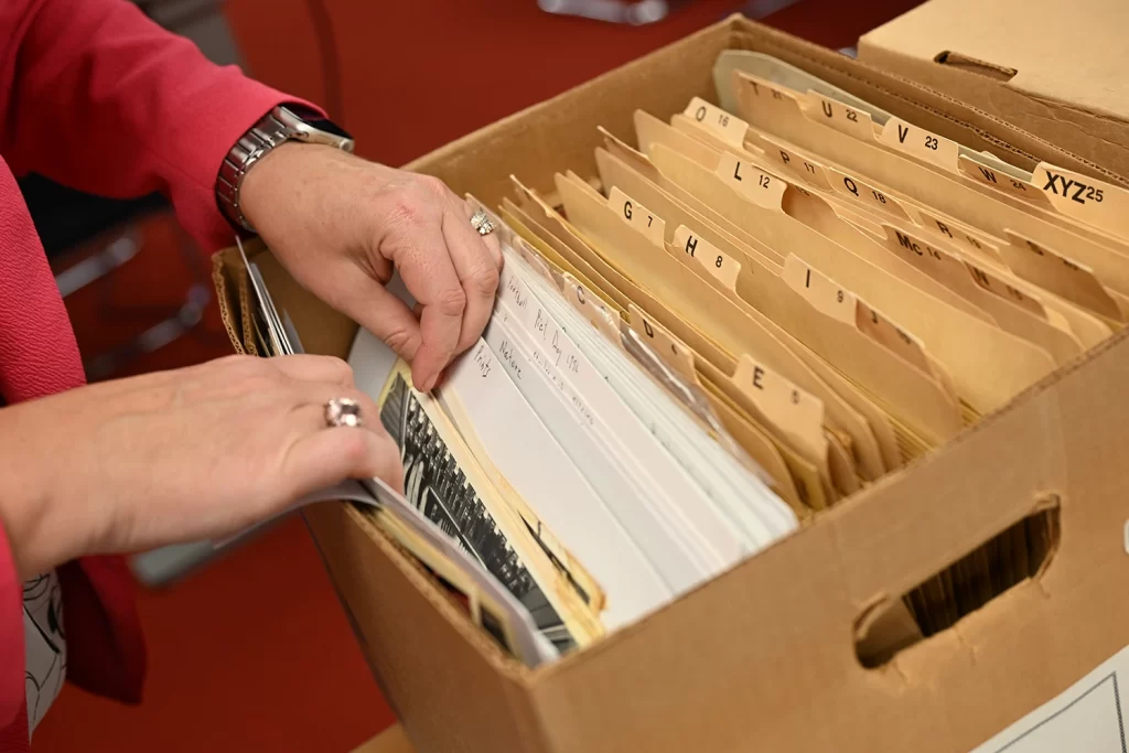 An open box on a table contains folders of organized papers from the Bentley archive. The hands of a user wearing a pink jacket are visible sorting through the materials.