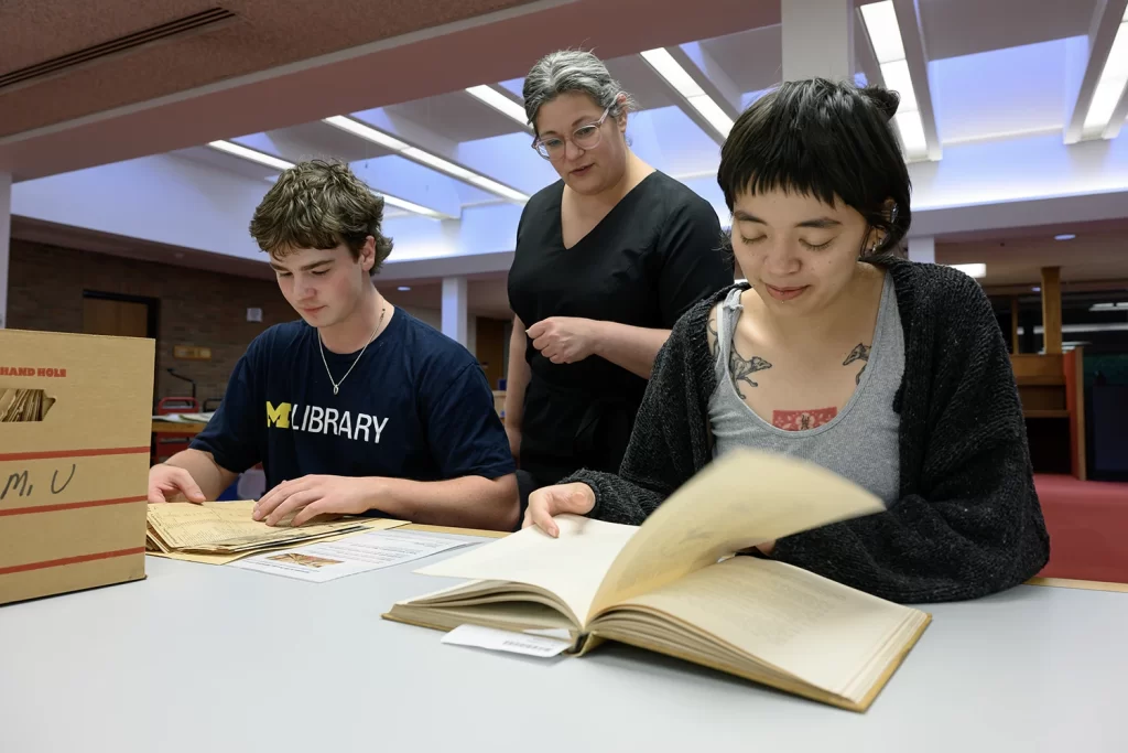Two researchers sit at a table looking down at historical papers. An archivist stands in the background looking over their shoulders.