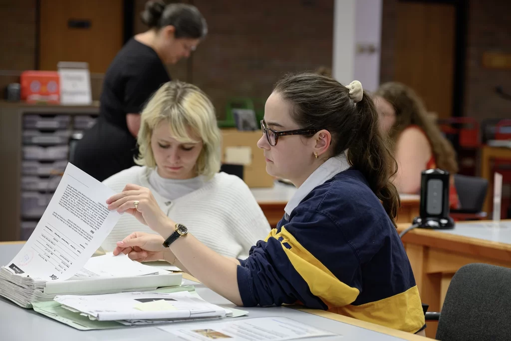 A woman wearing a maize and blue shirt studies a sheet of paper. Behind her another woman looks down at papers.
