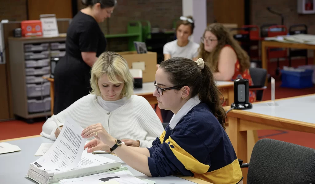 Visitors in the reading room of the Bentley Historical Library looking through historical documents at tables, featuring two researchers in the foreground, one wearing a striped shirt and glasses with her brown hair up in a ponytail, and one wearing a white sweater with bleached-blond hair, looking at documents together at one table. In the background, out of focus, an archivist in a black dress can be seen visiting another table with two researchers who have an open box of historical papers on the table in front of them.