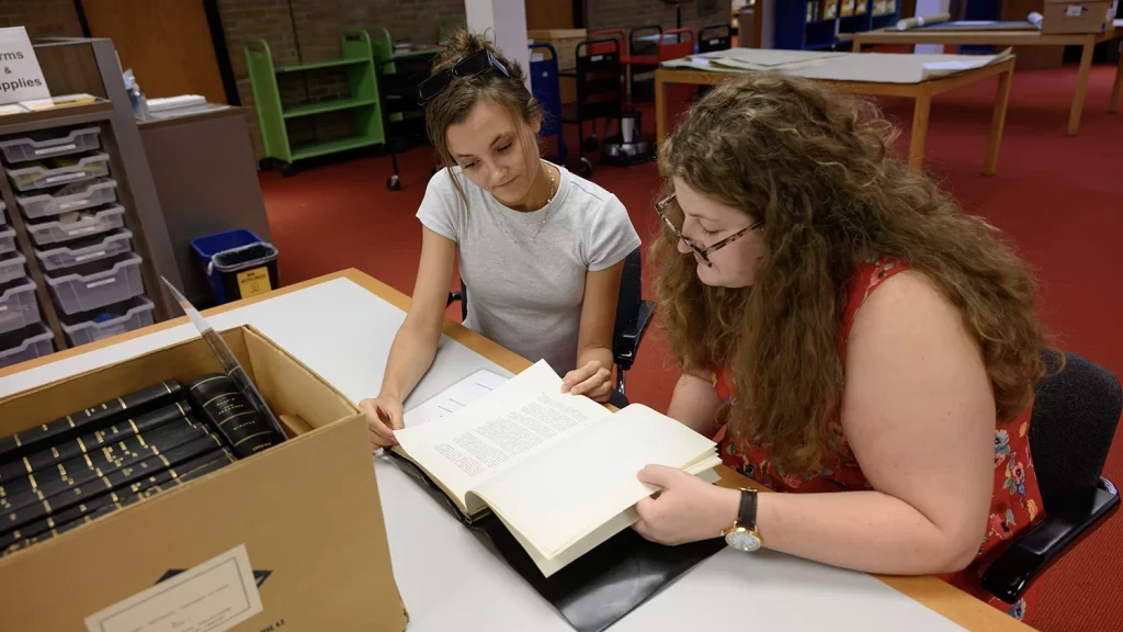 Two researchers at a table in the reading room looking at a historical book together. In front of them is a box of similar books, bound in black leather with gilt stamping. In the background are library carts and more tables.