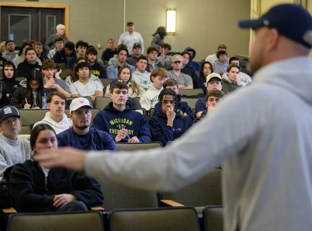 Professor Greg Dooley teaching the History of Athletics class at the University of Michigan. Professor Dooley is out of focus in the foreground, inviting you to focus on the audience full of U-M students, wearing t-shirts and U-M sweatshirts. Some students are looking at Professor Dooley, and some are taking notes.