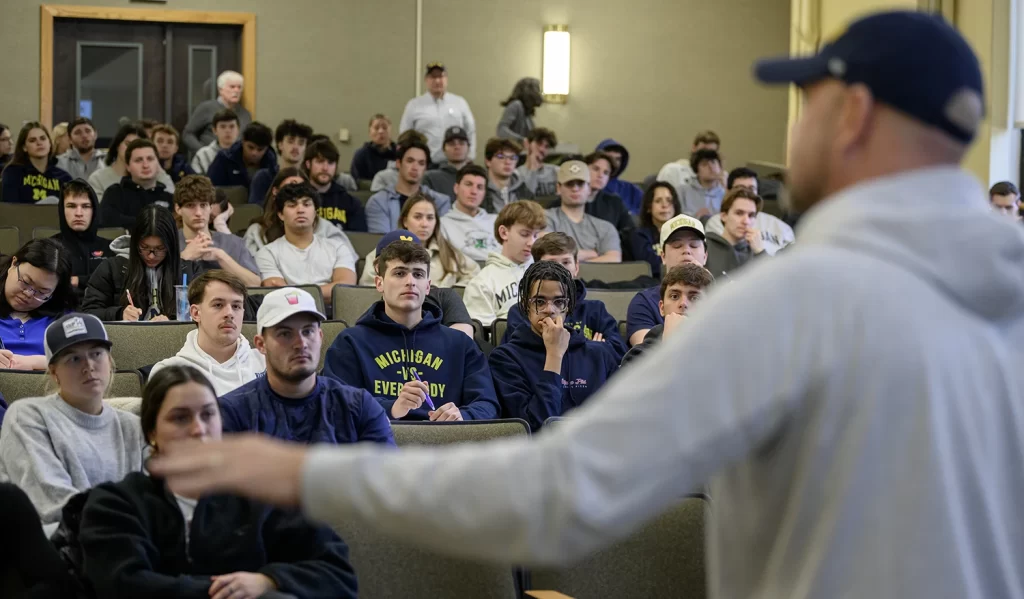 Professor Greg Dooley teaching the History of Athletics class at the University of Michigan. Professor Dooley is out of focus in the foreground, inviting you to focus on the audience full of U-M students, wearing t-shirts and U-M sweatshirts. Some students are looking at Professor Dooley, and some are taking notes.