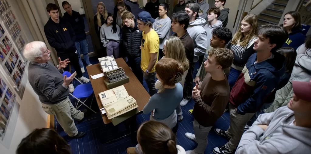 The University of Michigan's Athletics History class listening to Archivist Greg Kinney talk about the history of athletics at U-M, featuring a crowd of students standing around a table where a historical scrapbook is displayed, while Greg Kinney talks about U-M history.