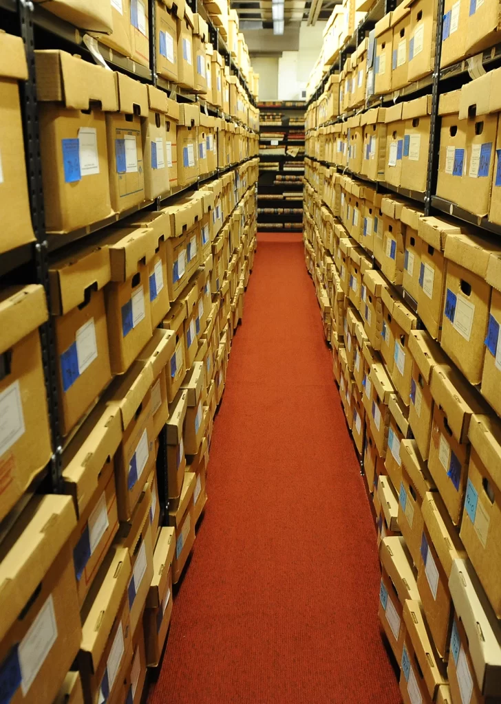 A long aisle with red carped is surrounded on both sides by tall shelves holding brown archival boxes.