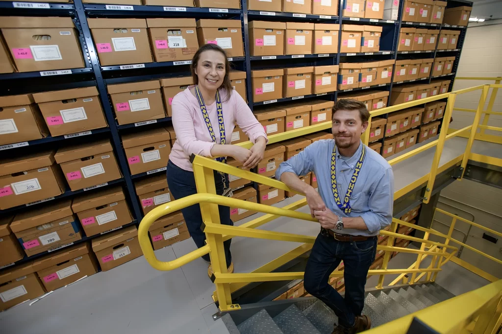 A male archivist stands on yellow stairs, while a female archivist stands near the stair railing. Behind them are a wall of boxes from the Bentley's collections.