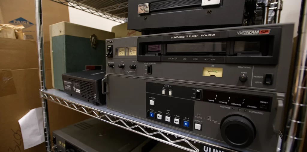 A large videocassette player stored on a white metal shelf alongside other machines used to play historical media like tapes.