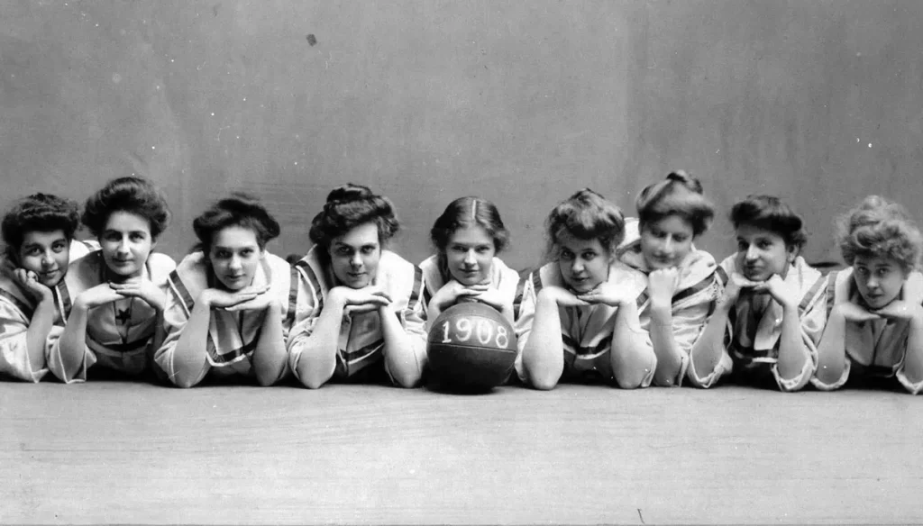 Nine women lay in a row with their chins on their hands in a black and white photo. In front of the center woman is a basketball with 1908 written on it.