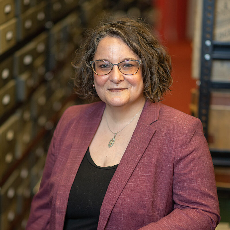 Bentley Director Alexis Antracoli stands in the Bentley Library stacks, with boxes and books in the background. She is wearing a mauve suit jacket and has dark curly hair and glasses.