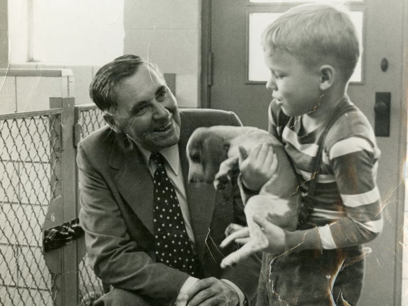 A man in a suit and tie crouching to smile at a boy in a striped shirt holding a puppy.