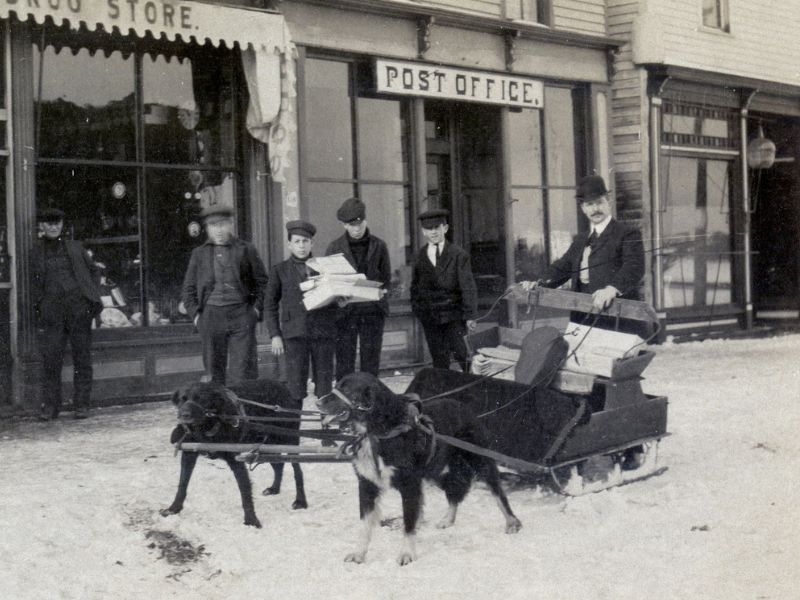 Two dogs pulling a mail sled, posed with four men in front of a Post Office in the snow.