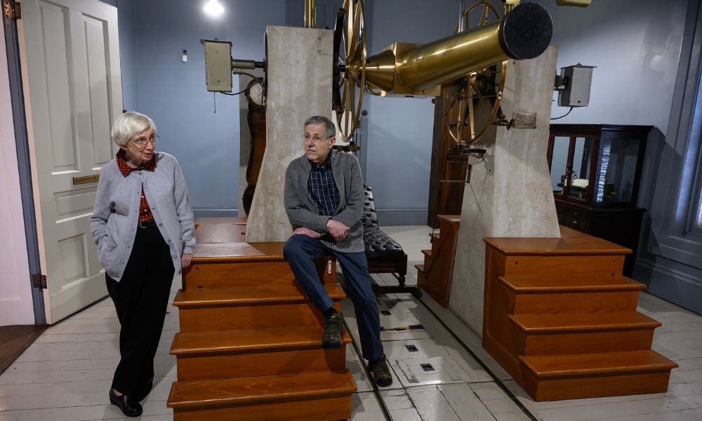 The Stenecks visiting the Detroit Observatory in the modern day, featuring Nick and Peg Steneck leaning against the steps of a historical telescope inside the Detroit Observatory.