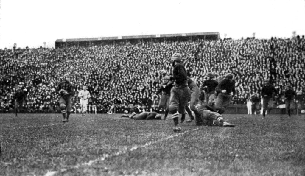 A black and white photo from 1919 of the Ohio State quarterback holding the football and the play developing in front of him.