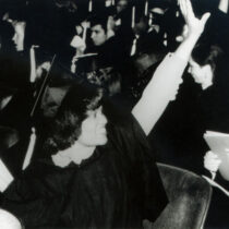 A woman wearing a graduation cap and gown with her hand in the air as if waving.