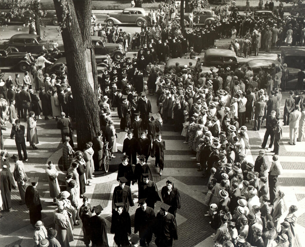 A black and white photo of many students in black graduation robes walking along a sidewalk. Spectators are lined up on each side to watch. 