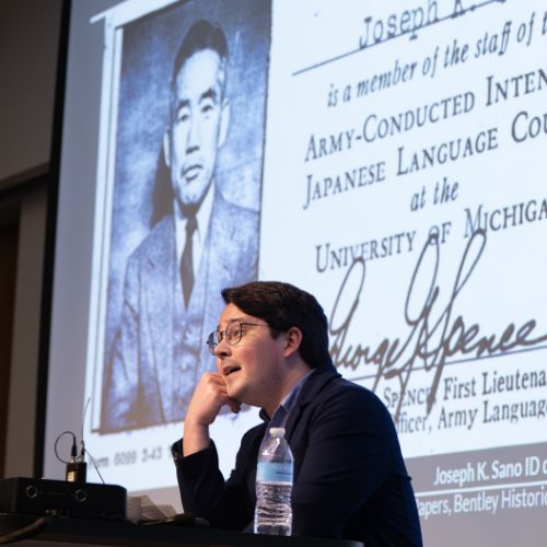 Researcher Brad Hammond stands at a podium at a Center for Japanese Studies event with an image of a Japanese American forcibly incarcerated during WWII on a screen behind him.