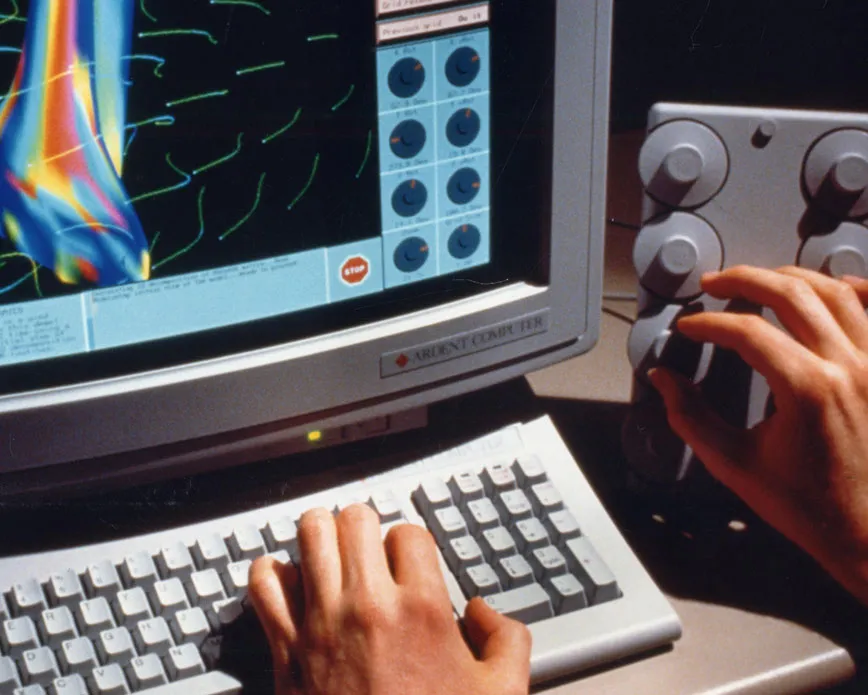A computer monitor showing graphics on the screen and a keyboard with a pair of hands manipulating keys and buttons.