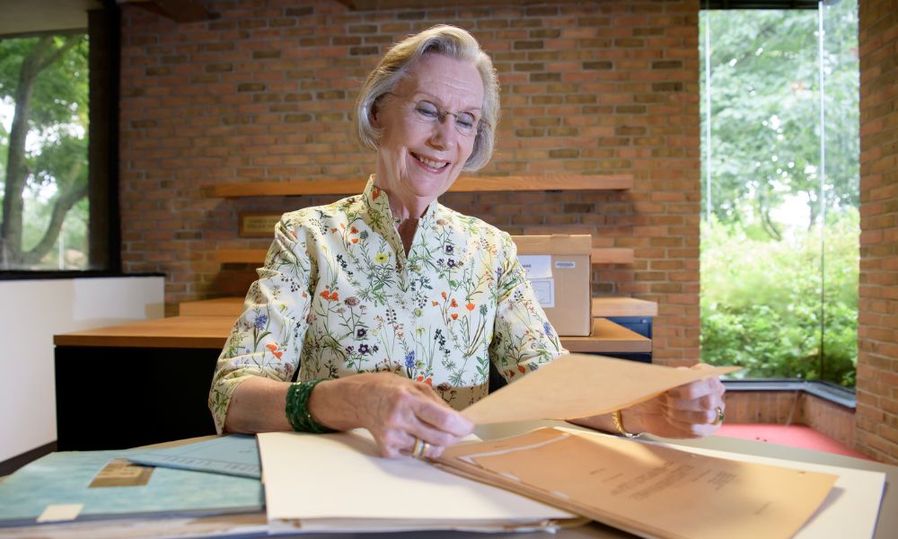 Alyce Sigler looking at historical documents inside the Bentley Historical Library, sitting at a table with historical papers on top of it, wearing a floral shirt, with a brick wall and a window showing greenery behind her.