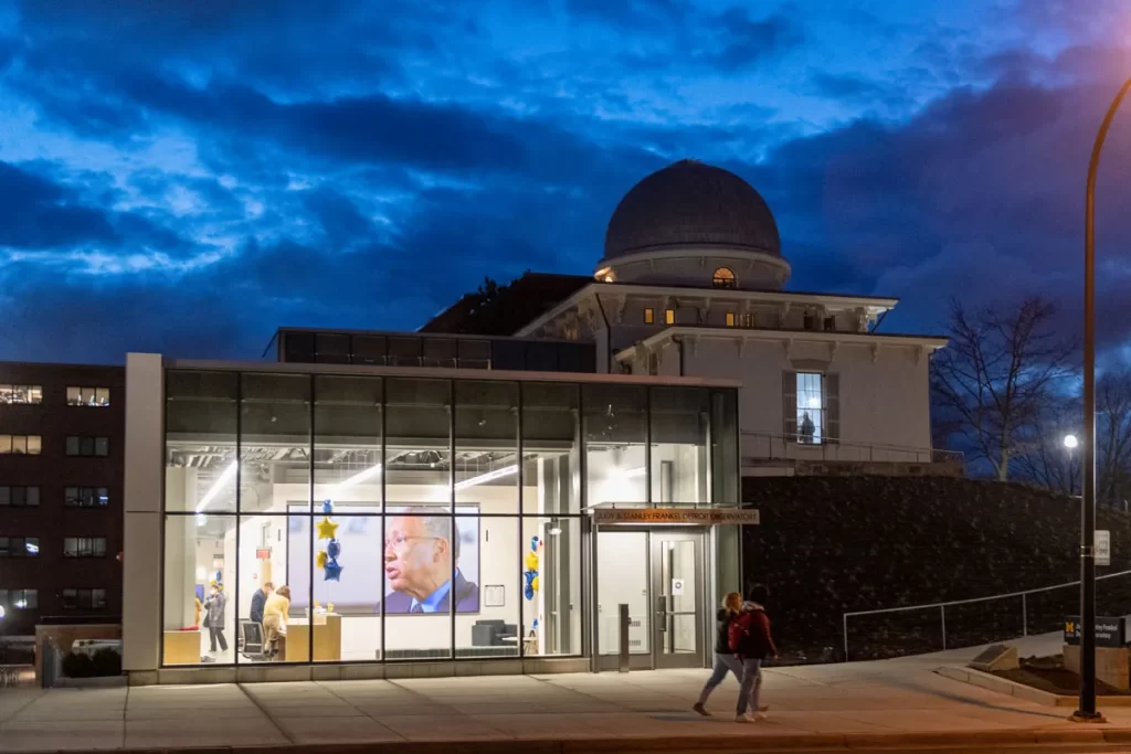 The Judy & Stanley Frankel Detroit Observatory building, featuring a historical building with an observatory dome, and the newer addition with glass paneling, illuminated at night, on U-M's Central Campus in Ann Arbor.