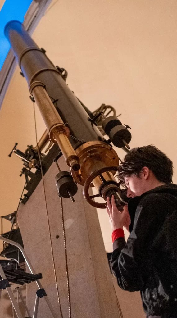 A man with dark hair wearing a dark shirt looks through a large historic telescope that is pointed up at the ceiling.