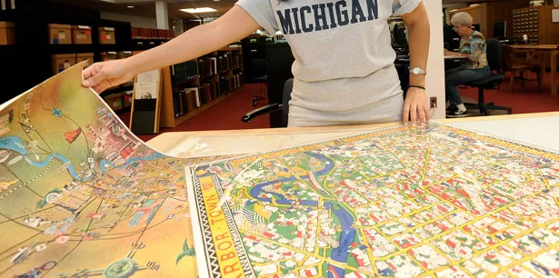 A person wearing a Michigan shirt uses both hands to spread out two large, colorful maps.