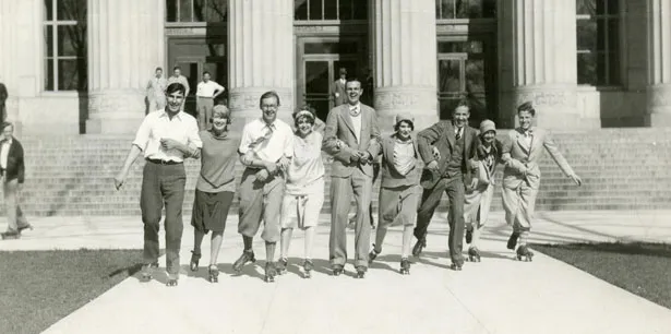 A black and white historical photo with nine University of Michigan students linking arms and rollerskating in front of a large building with pillars.