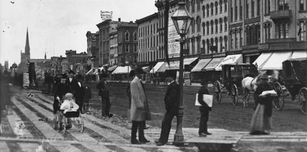 A black and white image of Woodward Ave. in Detroit circa the late 1800s, when it was a dirt road with horse-drawn carriages.