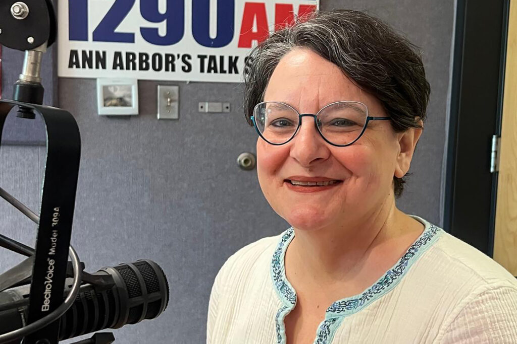 A woman with dark hair and glasses and a light-colored shirt sits in front of a podcast microphone.