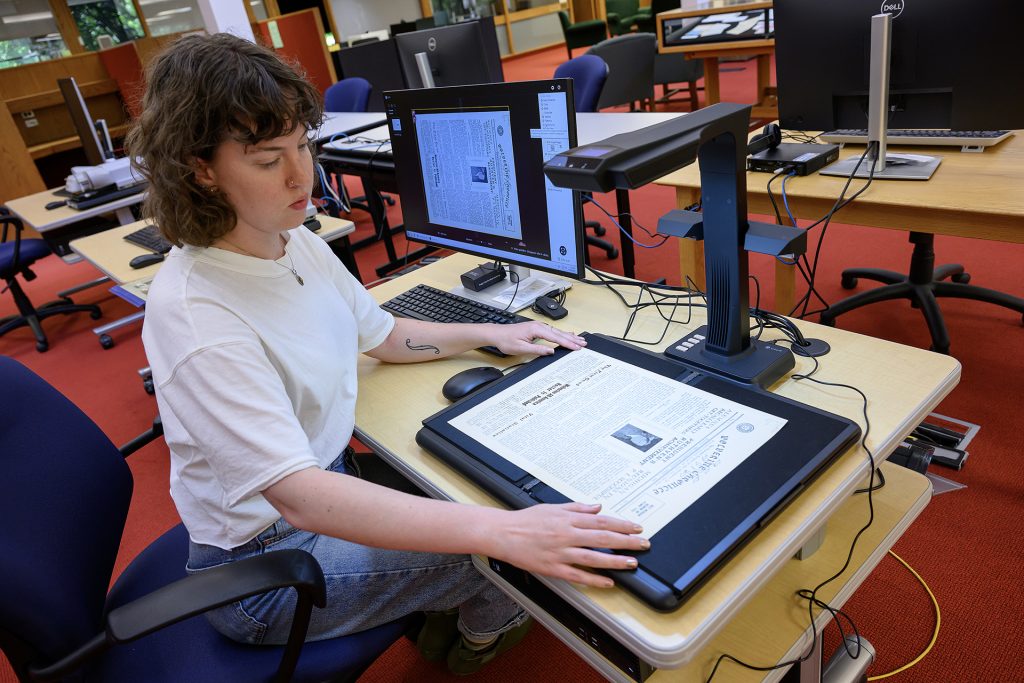 A woman with brown hair wearing a white shirt sits at a desk and adjusts a piece of paper on top of a black scanner bed. Behind her is a computer monitor displaying the paper on the scanner.