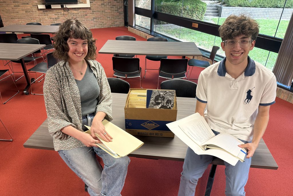 A man and woman lean against a table, each of them holding papers. The woman is on the left wearing a sweater and has brown hair, the man is on the right wearing a white shirt and has brown hair. A box of archival materials sits between them.