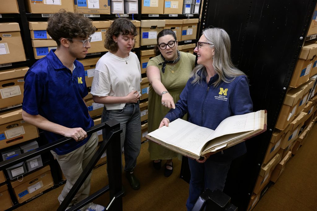 A woman wearing a blue jacket holds an oversized book. Three people on the left side of the image listen to her, one of them is pointing at the book. They are surrounded by boxes on shelves in an archive.