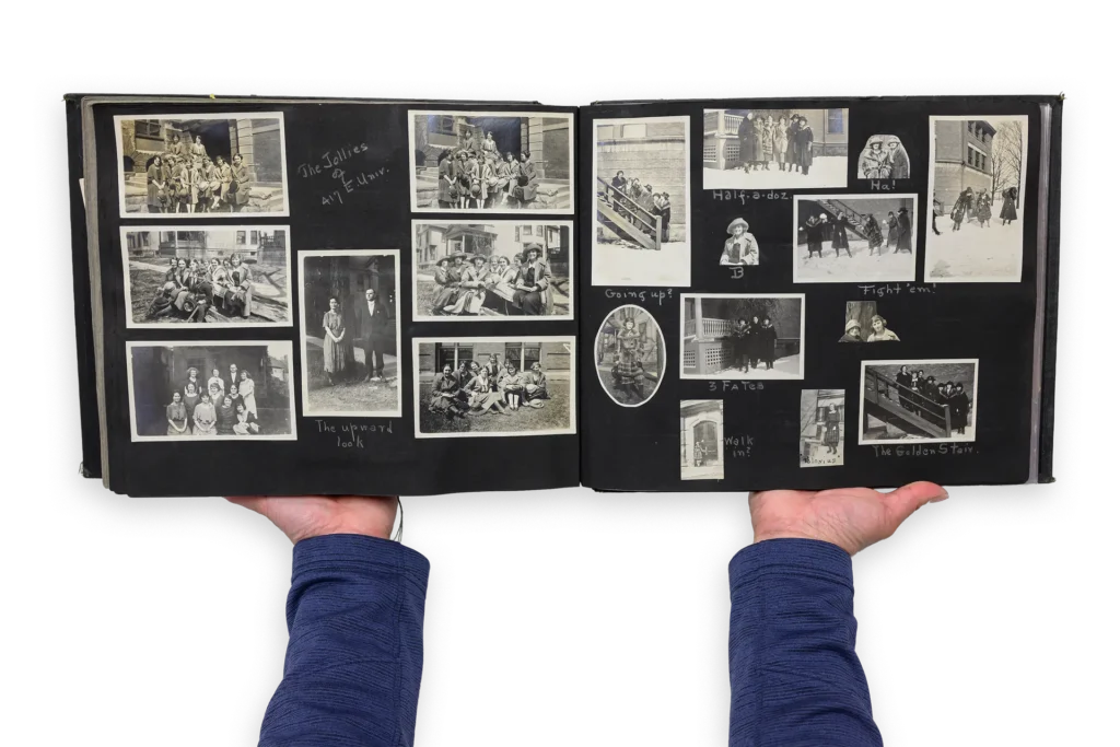 The open pages of a scrapbook show a dark background with many black and white photos pasted across the spread. Two hands hold the scrapbook open, and the arms are covered by dark blue sleeves.