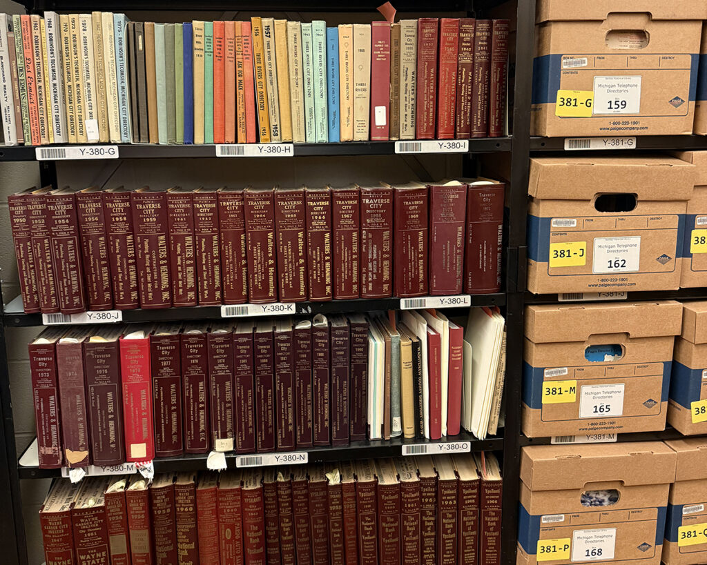 Black shelves with books on one side and brown archival boxes on the other, all stacked from floor to ceiling.