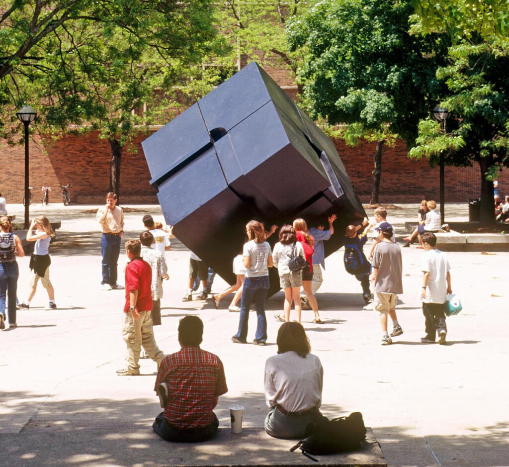 People surround a black cube sculpture on the University of Michigan Diag. Two people sit in the foreground with their backs to the camera.