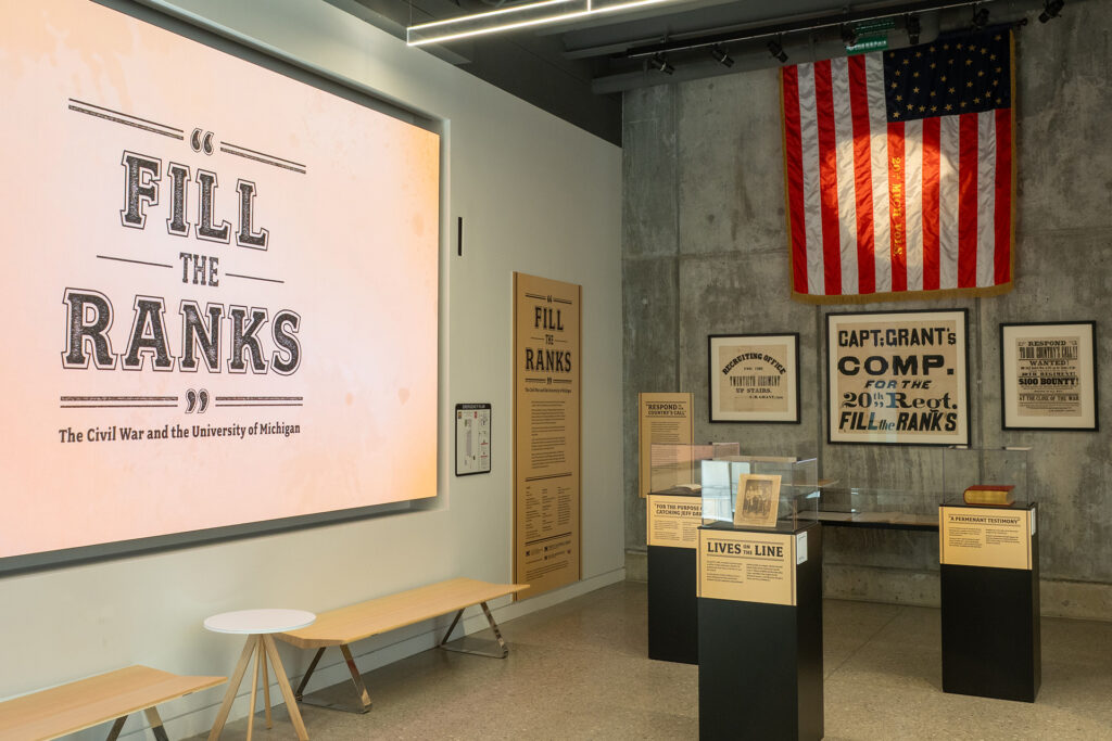 Exhibit cases in a grey-colored room with a digital screen on the wall that says "Fill the Ranks." A replica of a Civil War banner with red, white, and blue colors hangs on the back wall.