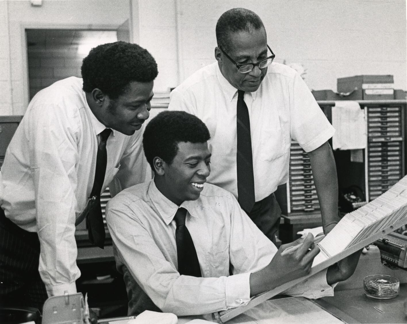 Photo of three Black men in white shirts and ties using card catalog.
