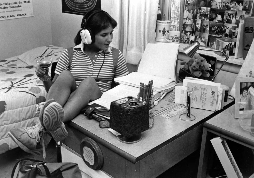 Photo of female student with headphones, reading book at desk in dorm room, 1980.