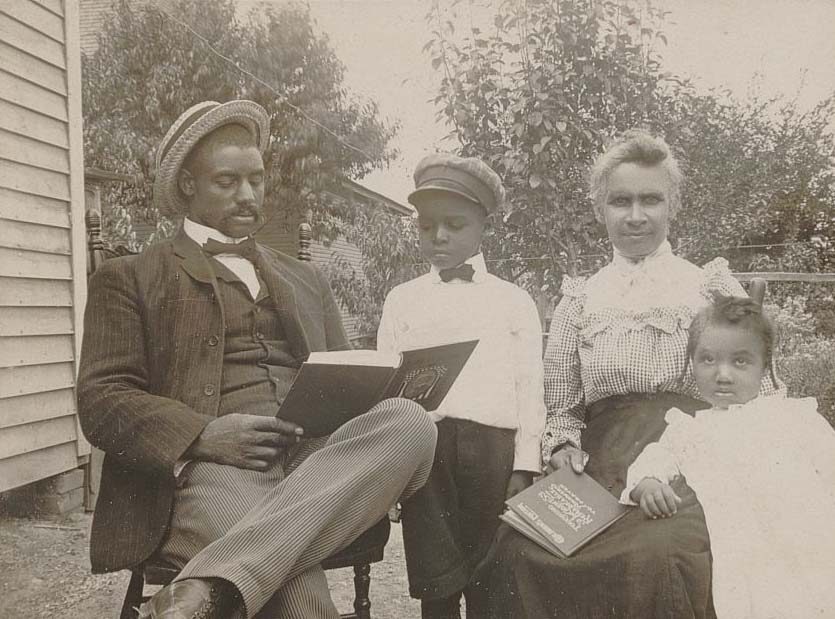 Outdoor family portrait - man and woman seated, holding books; boy reads over man